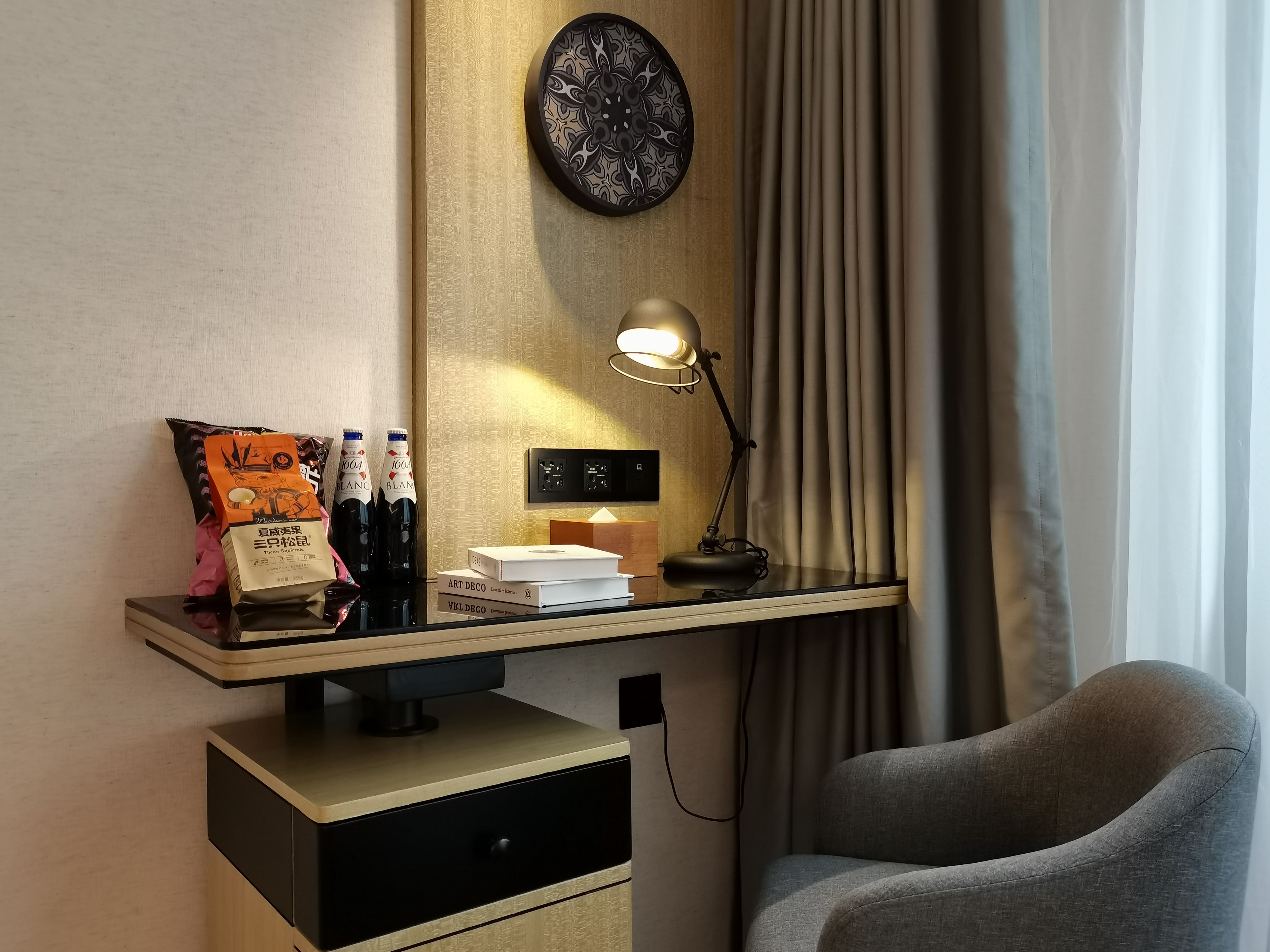 A hotel room with a desk, a lamp, a chair, and a wall clock.