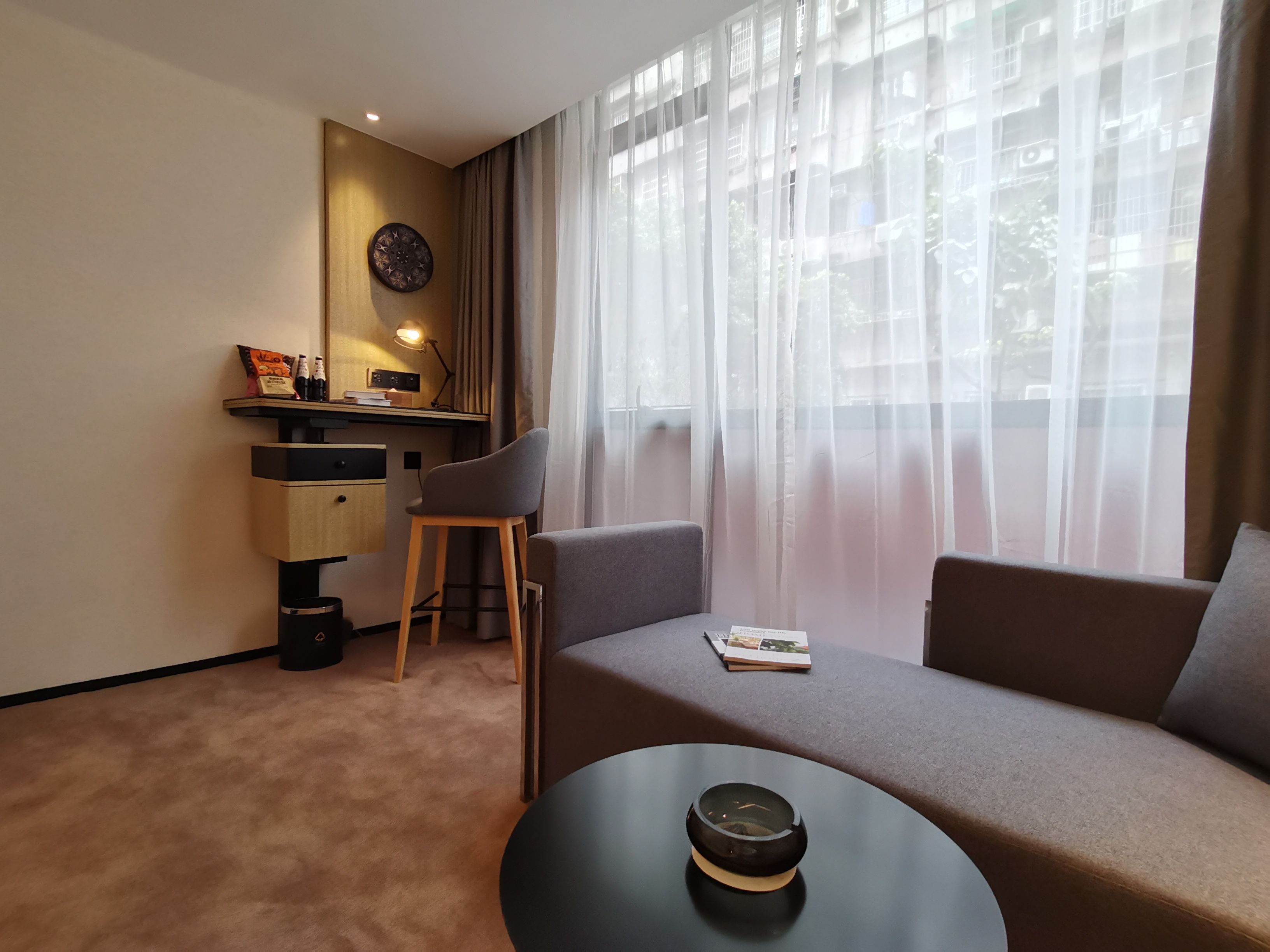 A modern living room with a sofa, a coffee table, a desk with a chair, and a large window with sheer curtains.