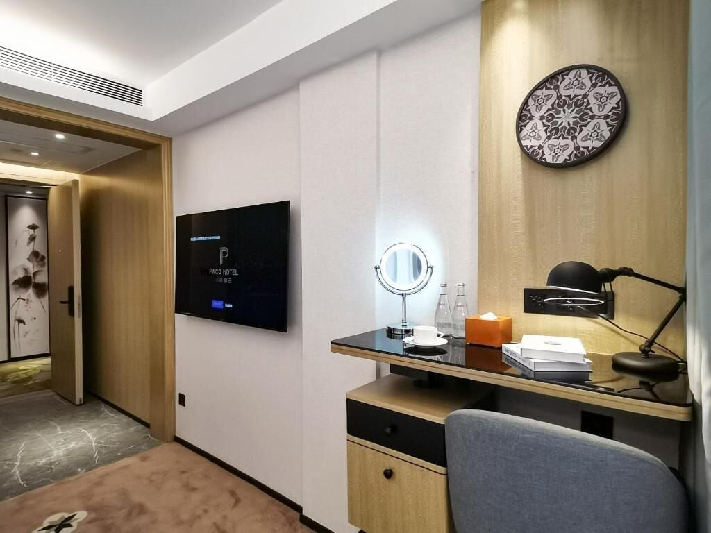A hotel room with a flat-screen TV mounted on the wall, a vanity with a mirror, a desk with a lamp, and a bed with a headboard.