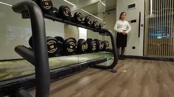 Une salle de sport avec une rangée d’haltères sur un support, une personne debout à côté du support, et une porte vitrée à l’arrière-plan.