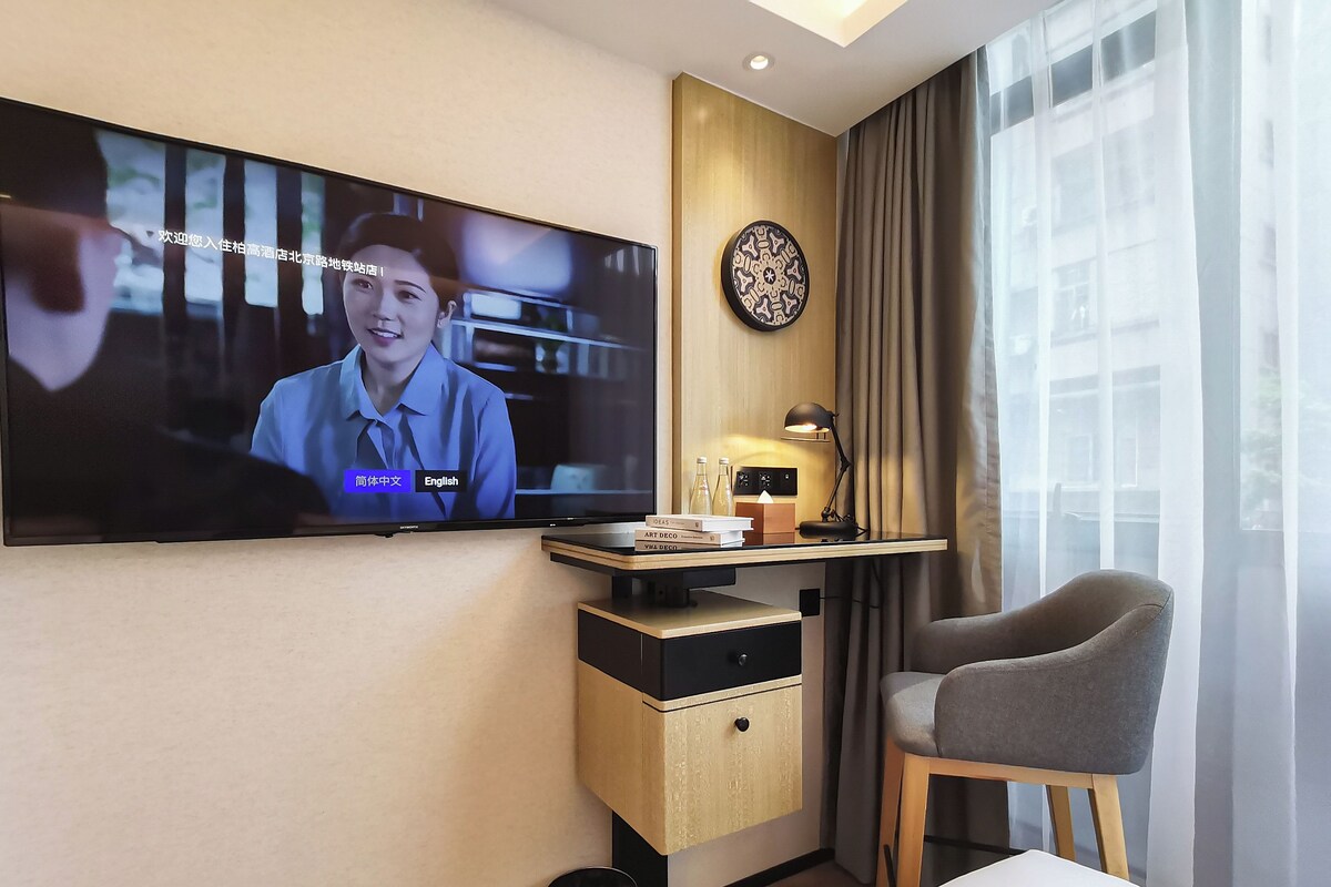 Habitación de hotel con televisión montada en la pared, un escritorio con lámpara, una silla y una mesita de noche.