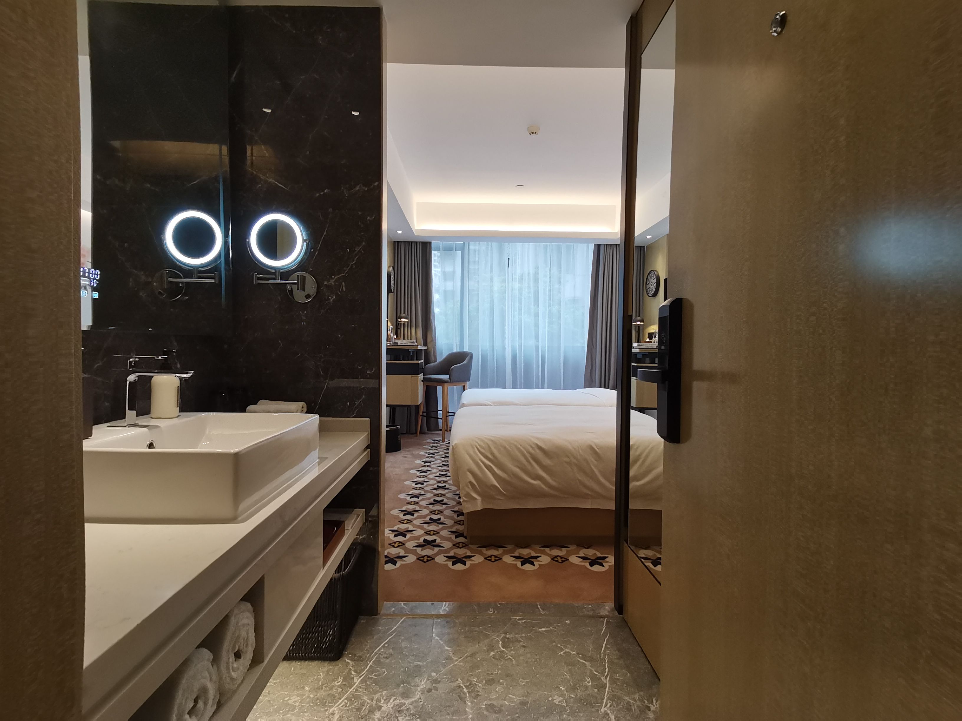 Een moderne hotelkamer met een groot bed, nachtkastje, bureau, stoel, een badkamer met spiegel en wastafel, en uitzicht op de omgeving.