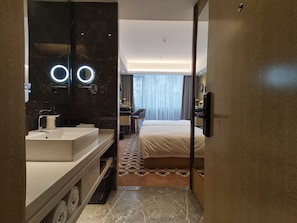 A modern hotel room with a large bed, a bedside table, a desk, a chair, a bathroom with a mirror and sink, and a view of the outdoors.