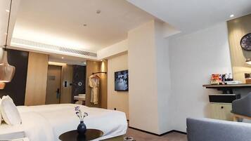 Quarto de hotel com cama, mesa de cabeceira, abajur e uma pequena mesa com um vaso. Há um sofá e uma escrivaninha com uma cadeira.