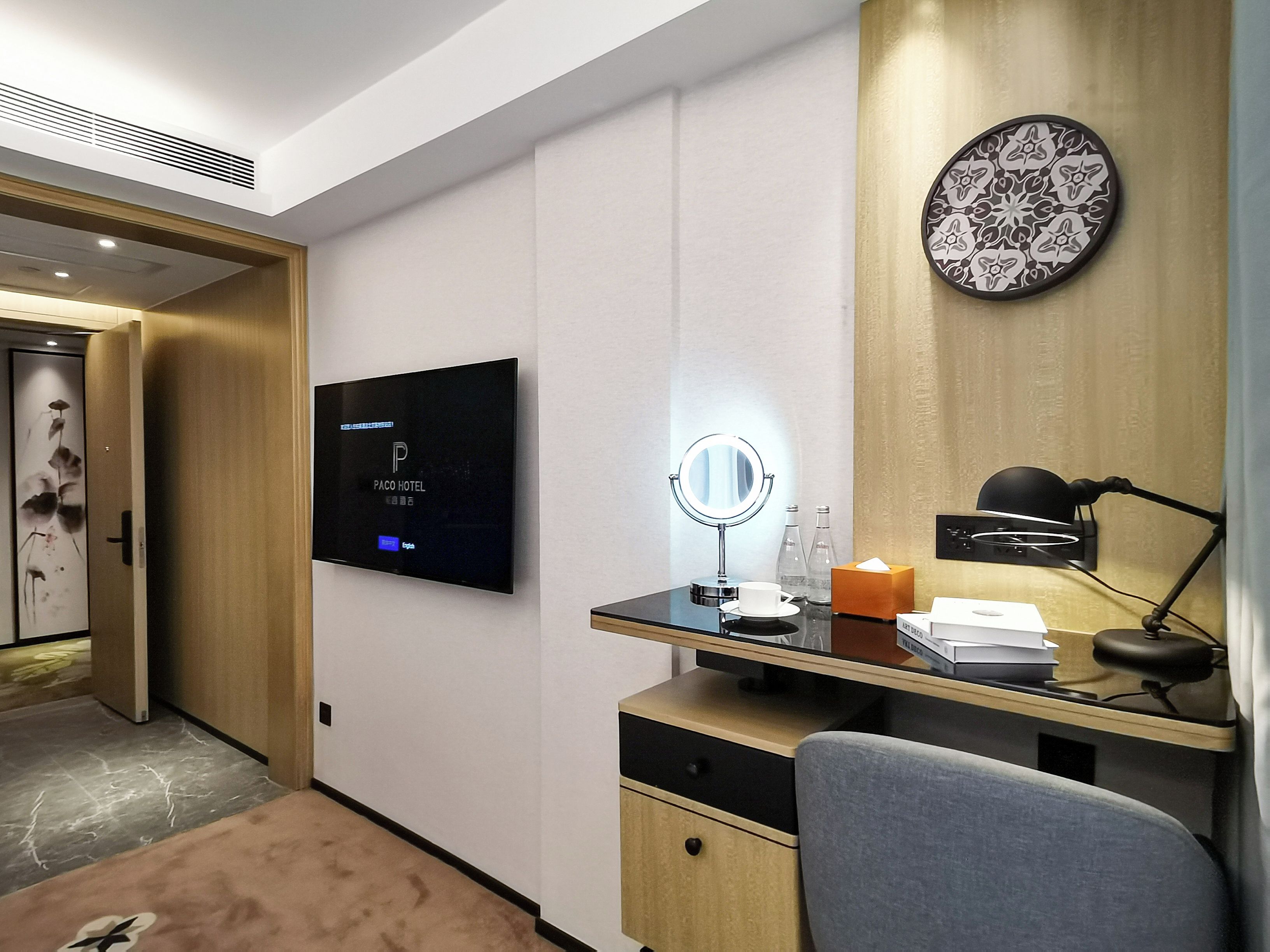 A modern hotel room with a flat-screen TV, a desk with a mirror, a lamp, and a decorative plate on the wall.