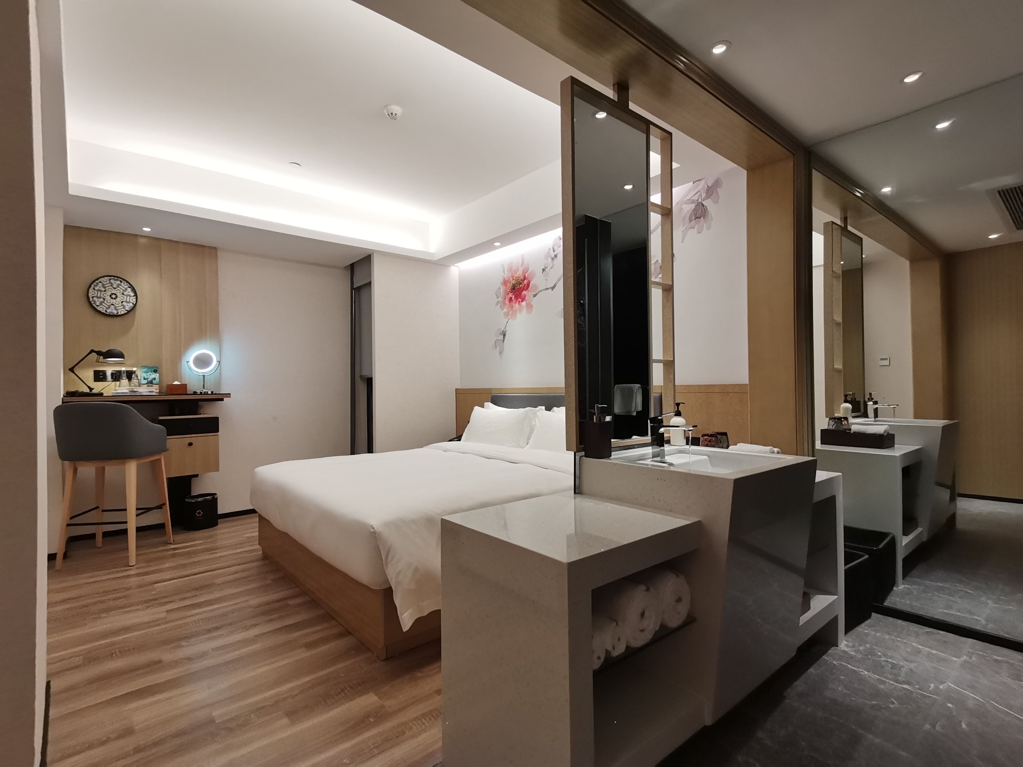 A modern hotel room with a bed, a desk with a chair, a large mirror, and a bathroom with a sink and mirror.