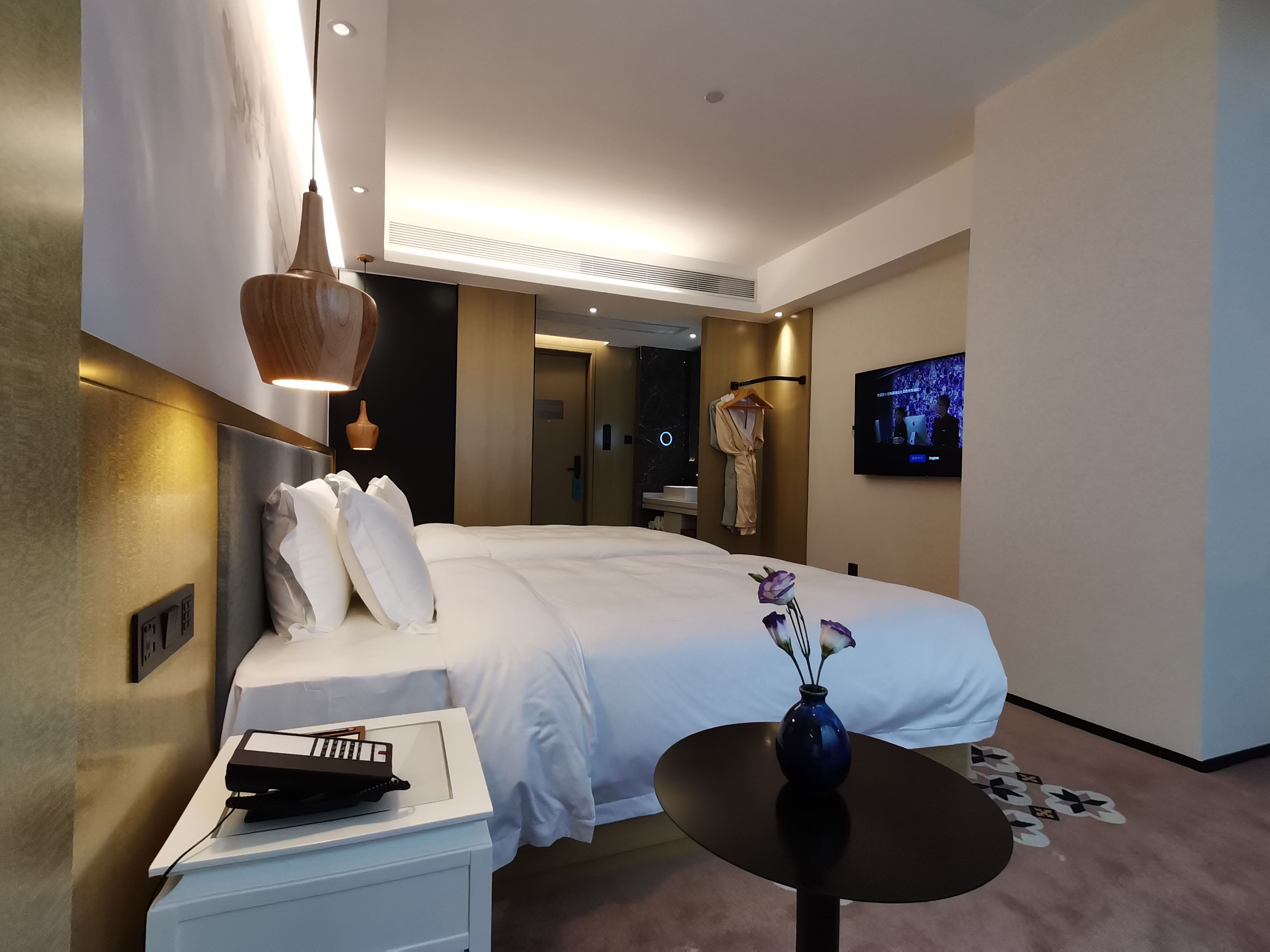 A modern hotel room with a neatly made bed, a bedside table with a remote control, a small table with a vase, and a television mounted on the wall.