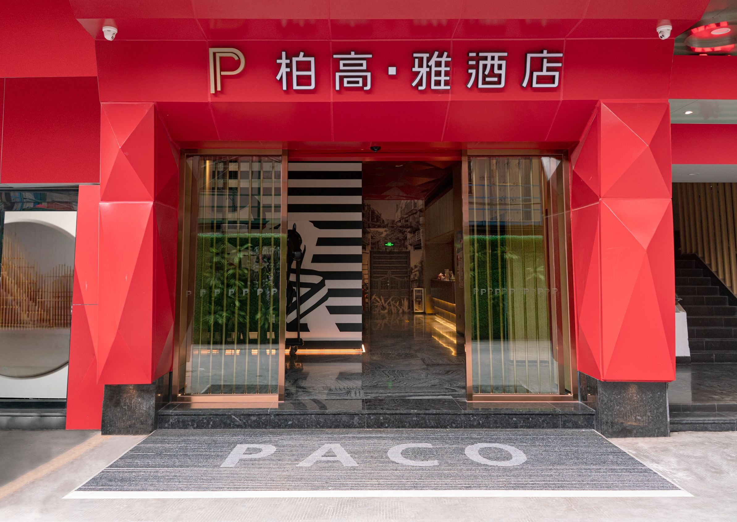 La entrada de un hotel con un llamativo letrero con la letra 'P' y un felpudo que dice 'PACO'.