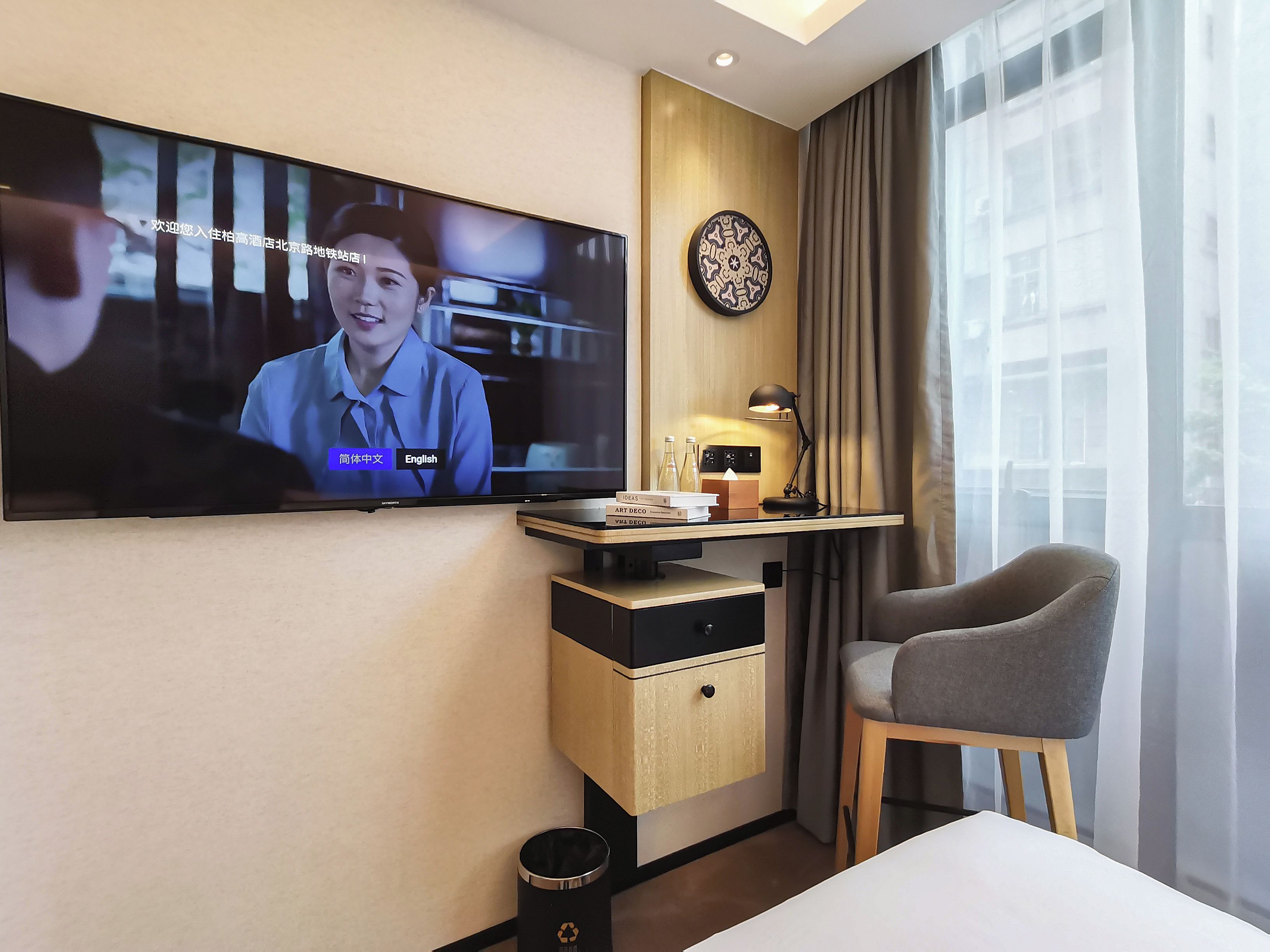 A hotel room with a television mounted on the wall, a desk with a lamp, a chair, and a bedside table.