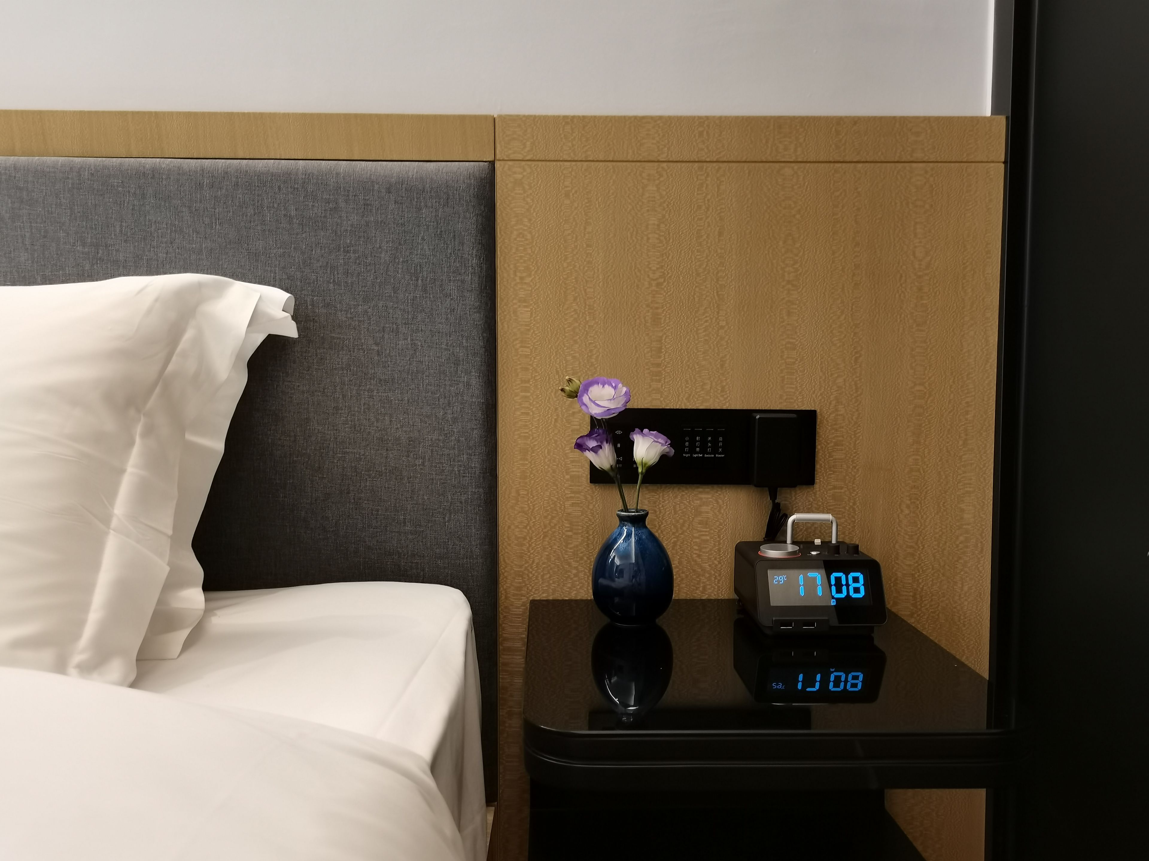 A hotel room with a bed, a nightstand with a digital clock, and a vase with flowers.
