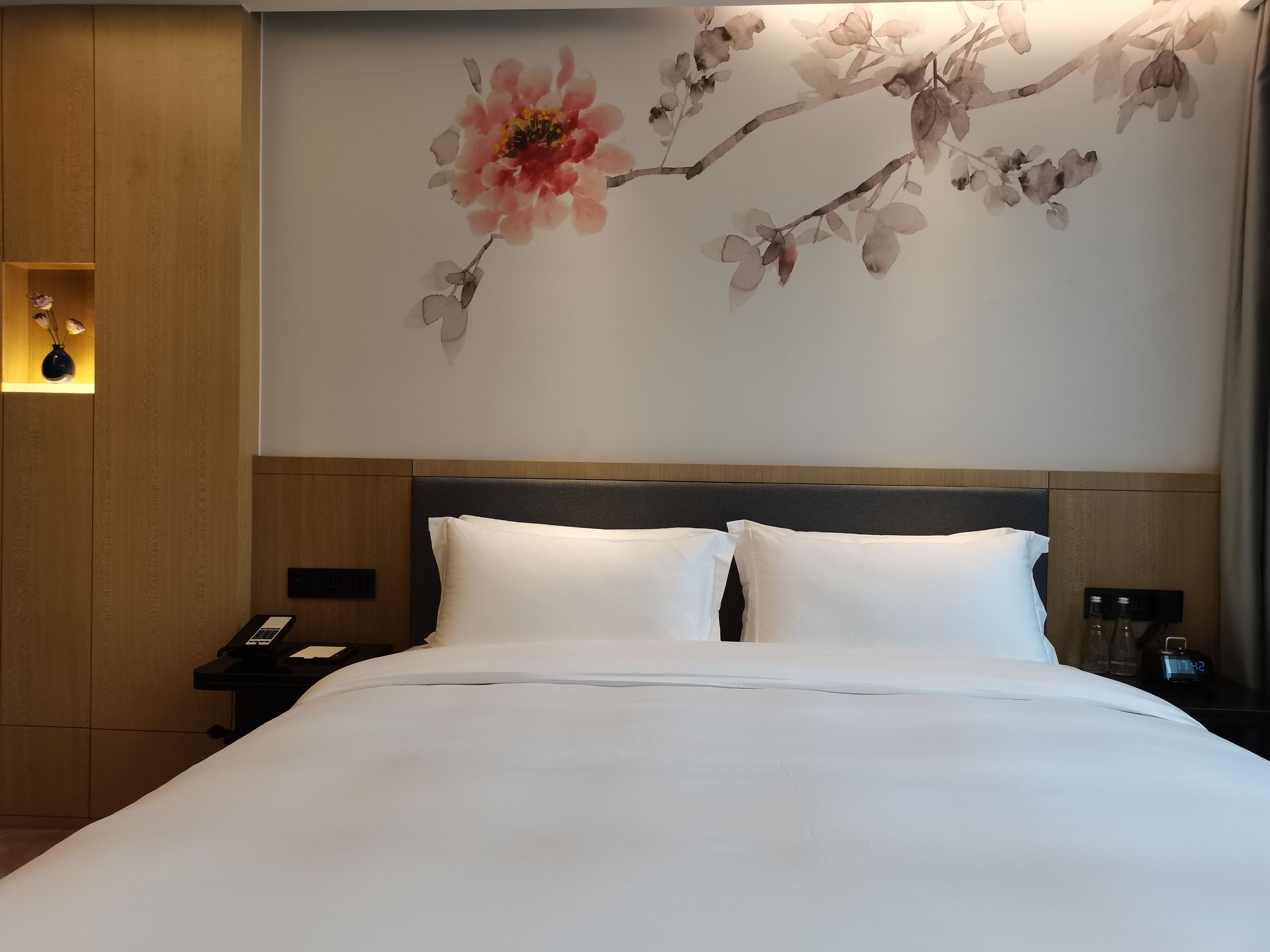 Ein Hotelzimmer mit einem Bett, einem Nachttisch, einer Wanduhr und einem floralen Kunstwerk.