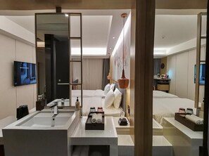 Habitación de hotel moderna con una cama grande, baño separado con un espejo amplio, televisión y mesita de noche con artículos de cortesía.