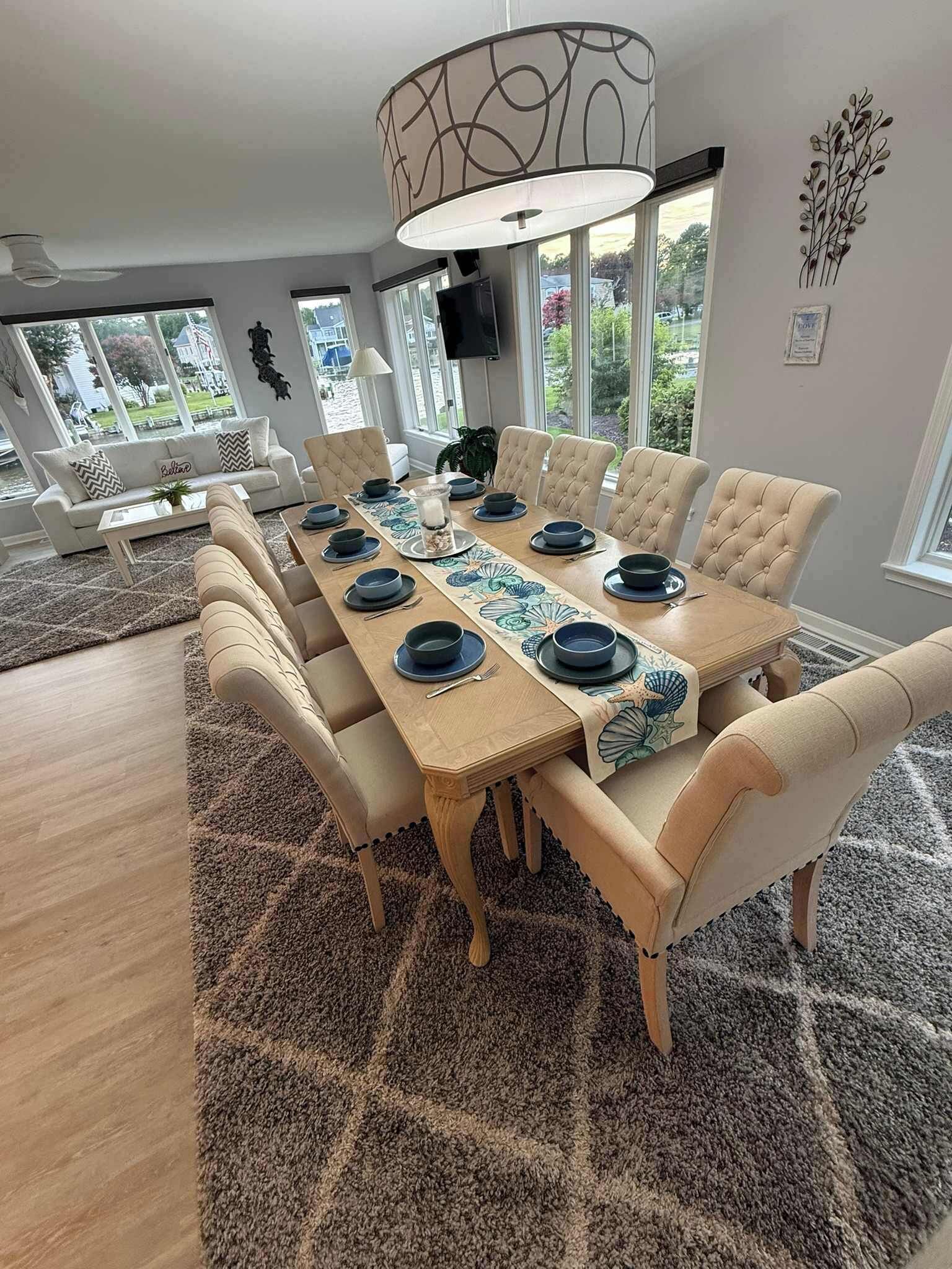 A dining room with a long table set for six, surrounded by cushioned chairs, a patterned table runner, and a large pendant light.