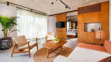 Una sala moderna con un sofá, un sillón y una mesa de centro. Hay un televisor montado en la pared y un ventanal con cortinas.