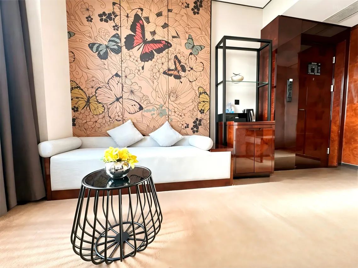 Een hotel lobby met een bank, een ronde tafel met een bloevas, een decoratieve wand met bloemenmotieven en een lift.