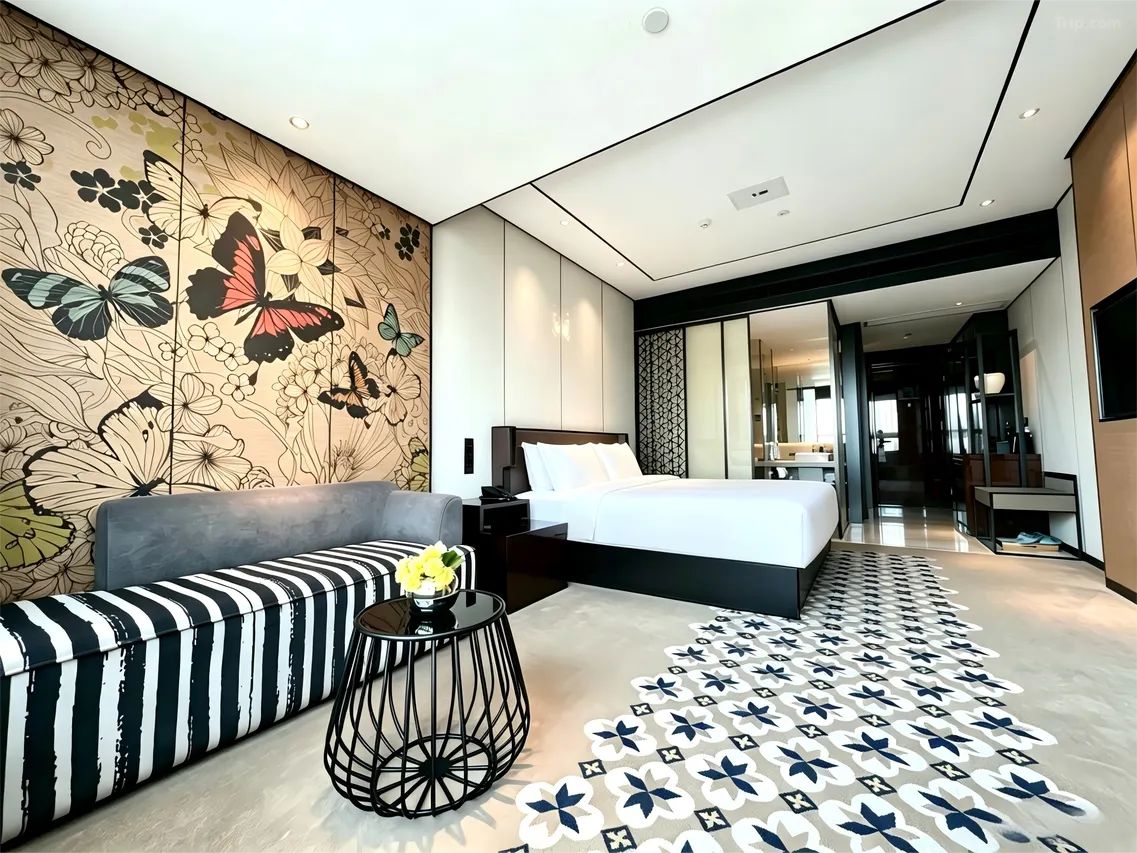 Une chambre d’hôtel moderne avec un grand lit, un canapé, une petite table ronde ornée d’un vase et un sol à motifs.