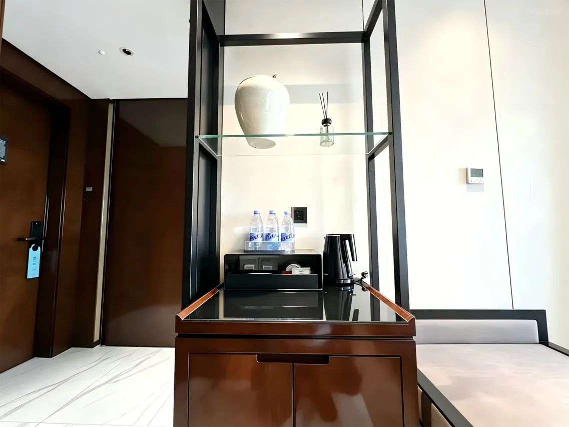 Een hotelkamer met een houten bureau, een glazen plankje met een vaas, een koffiezetapparaat en een minibar.