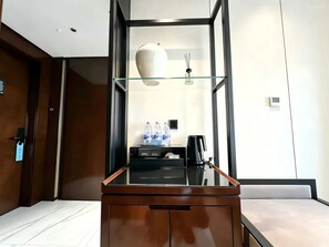 A hotel room with a wooden desk, a glass shelf with a vase, a coffee maker, and a minibar.