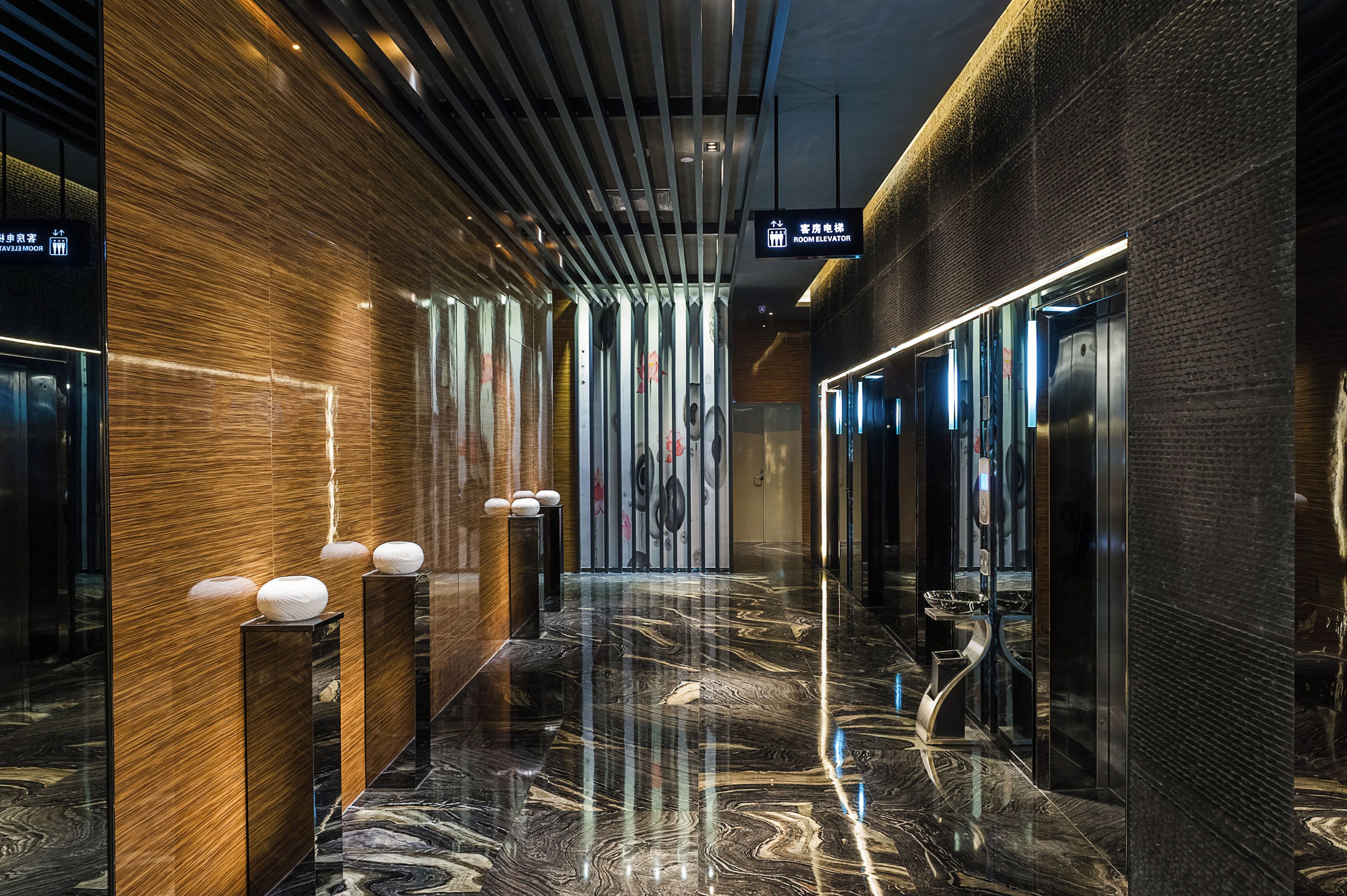 Um hall de hotel moderno com piso de mármore, detalhes em madeira e um elevador de vidro.