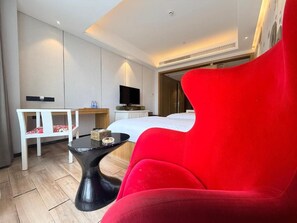 A modern hotel room with a red armchair, a bed, a desk, and a television.