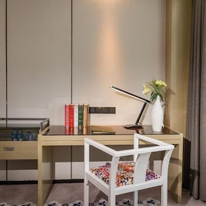 A wooden desk with a white chair, a vase with flowers, and a lamp. There are books on the desk and a small shelf with decorative items.