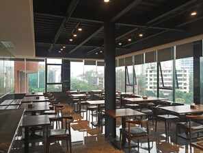Un espacio interior amplio con mesas y sillas dispuestas para comer, ventanas grandes con vistas a edificios y zonas verdes, y un techo alto con vigas e iluminación a la vista.
