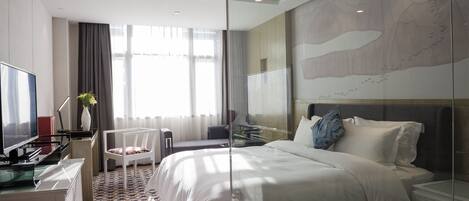 A modern hotel room with a large bed, a desk with a TV, and a glass partition.