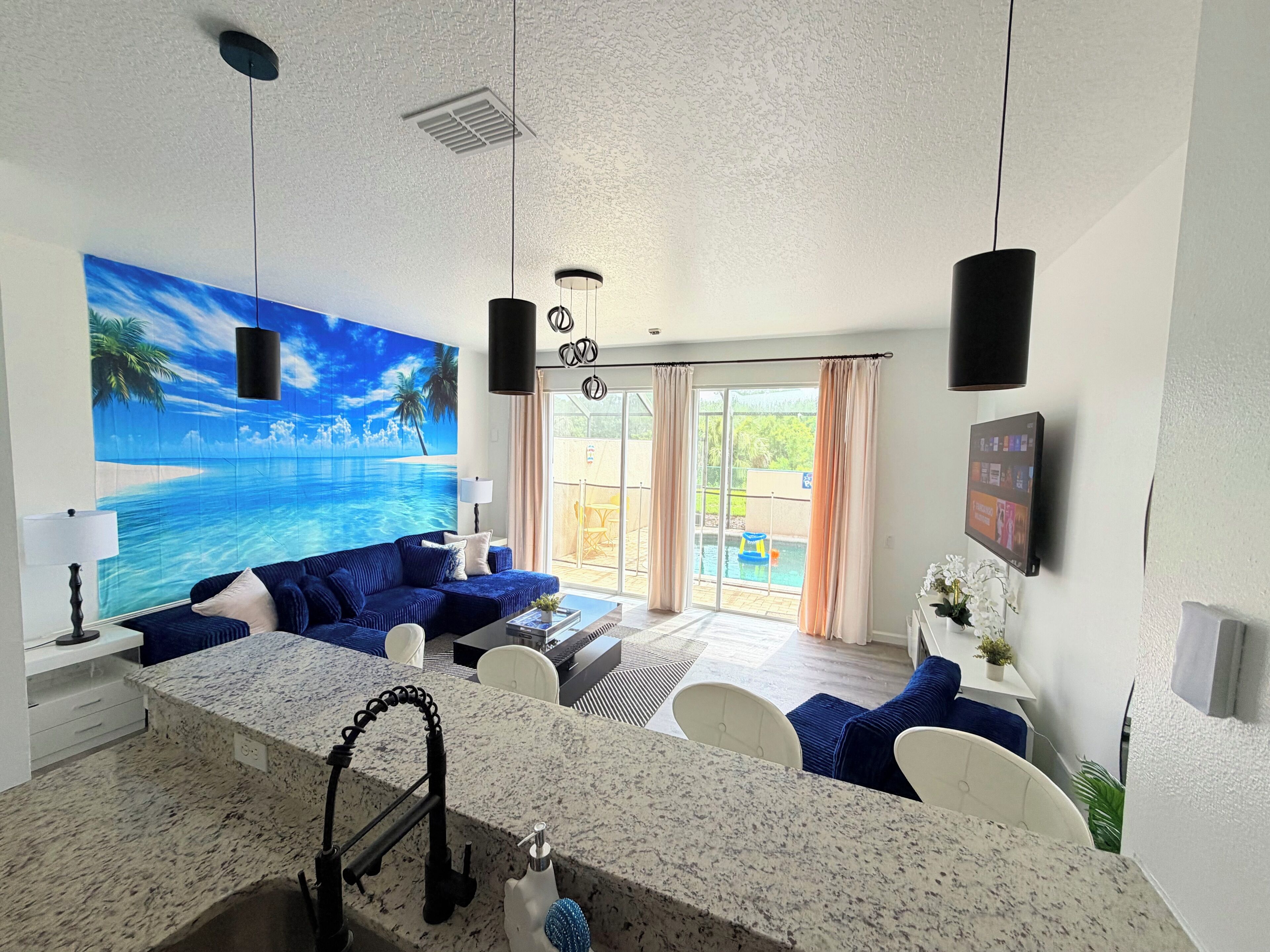 A modern living room with a large window, a blue sofa, a kitchen area with a granite countertop, and a ceiling fan.