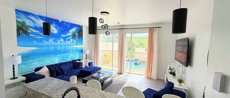 A modern living room with a large window, a blue sofa, a kitchen area with a granite countertop, and a ceiling fan.