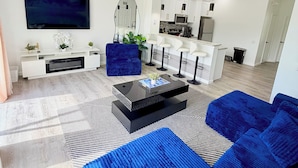 Een moderne woonkamer met een grote flatscreen televisie, een salontafel, een blauwe bank en een keuken op de achtergrond.