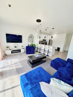 A modern living room with a large flat-screen TV, a coffee table, a blue sofa, and a kitchen in the background. - Disney Family Home/3-bedroom townhome with private pool in Kissimmee. (Kissimmee)
