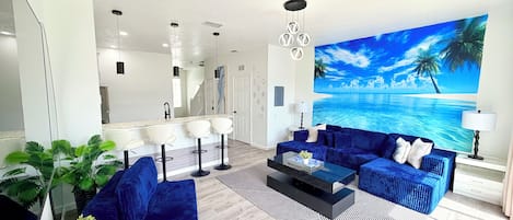 A modern living room with a blue sofa set, a coffee table, and a large tropical mural on the wall.