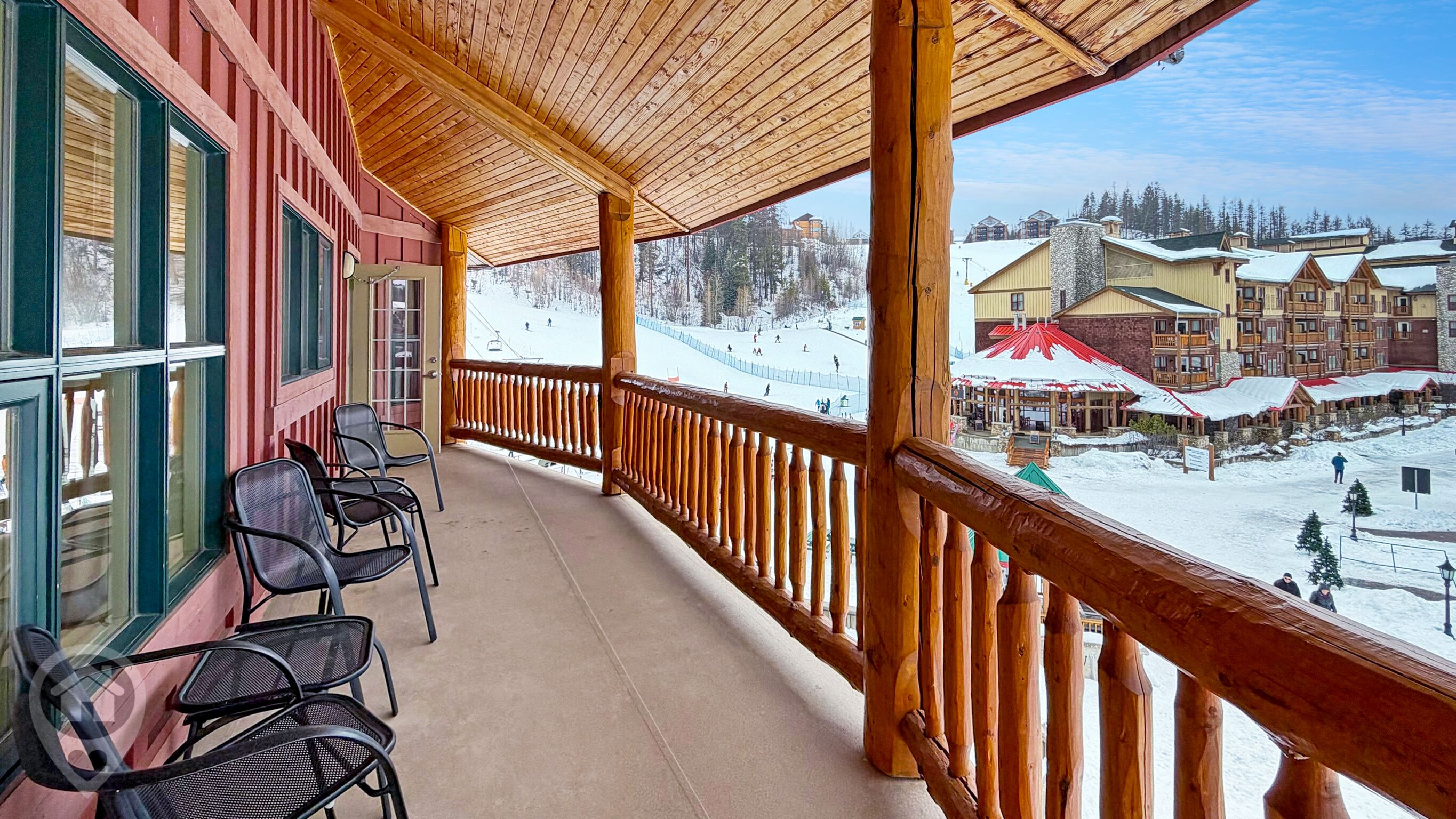 A wooden balcony with chairs, overlooking a snowy ski resort with buildings and a ski lift.