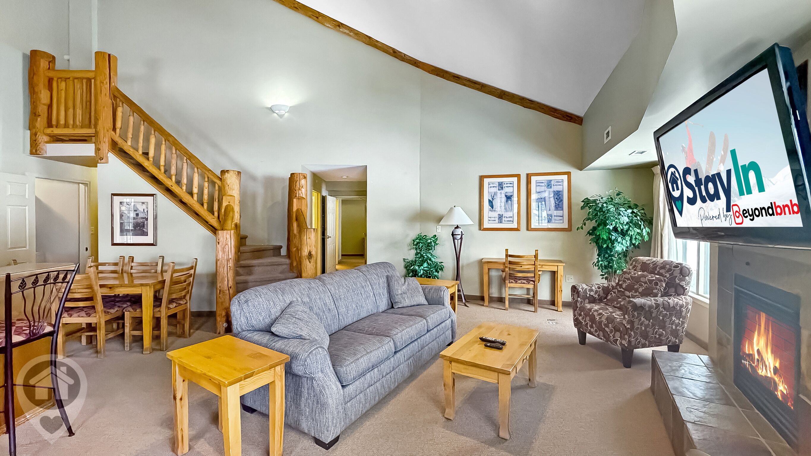 Ein geräumiges Wohnzimmer mit einer Holztreppe, einem Kamin und einem Fernseher, der 'Stay In' anzeigt.
