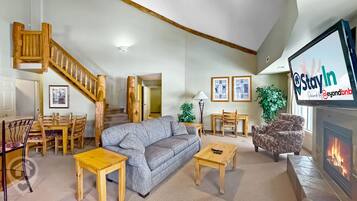 A spacious living room with a wooden staircase, a fireplace, and a television displaying 'Stay In'.