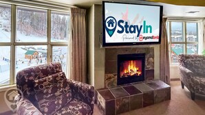 A living room with a fireplace, a television mounted above it, and large windows overlooking a snowy outdoor area.