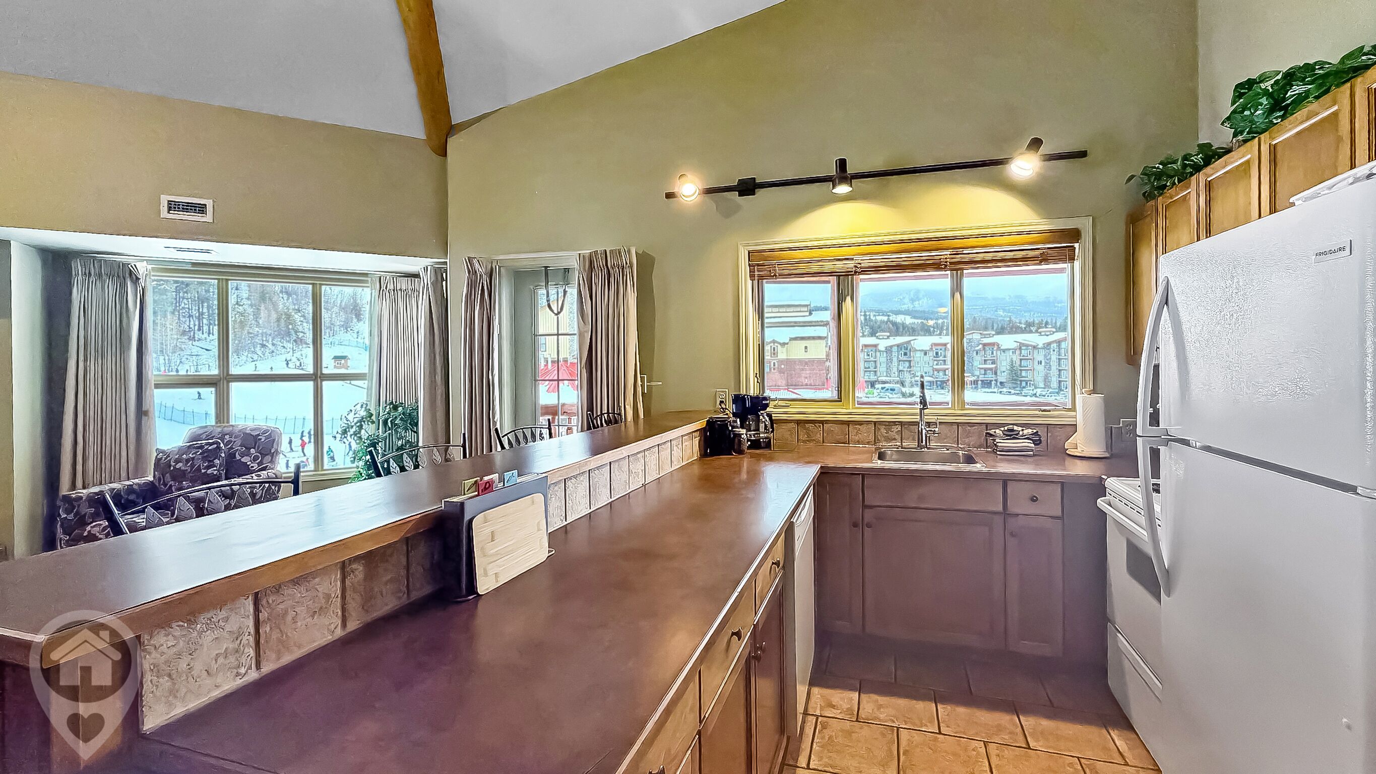 A kitchen with a long counter, a sink, and a refrigerator. There are windows with curtains, and a view of buildings outside.