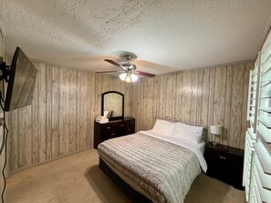 Un dormitorio con cama, cómoda, espejo, ventilador de techo y televisor montado en la pared.