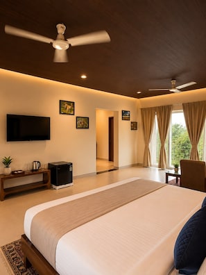 A modern bedroom with a large bed, ceiling fans, a TV, artwork, and a view of greenery.