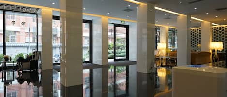 A modern hotel lobby with a reflective floor, large windows, and a seating area with a lamp.