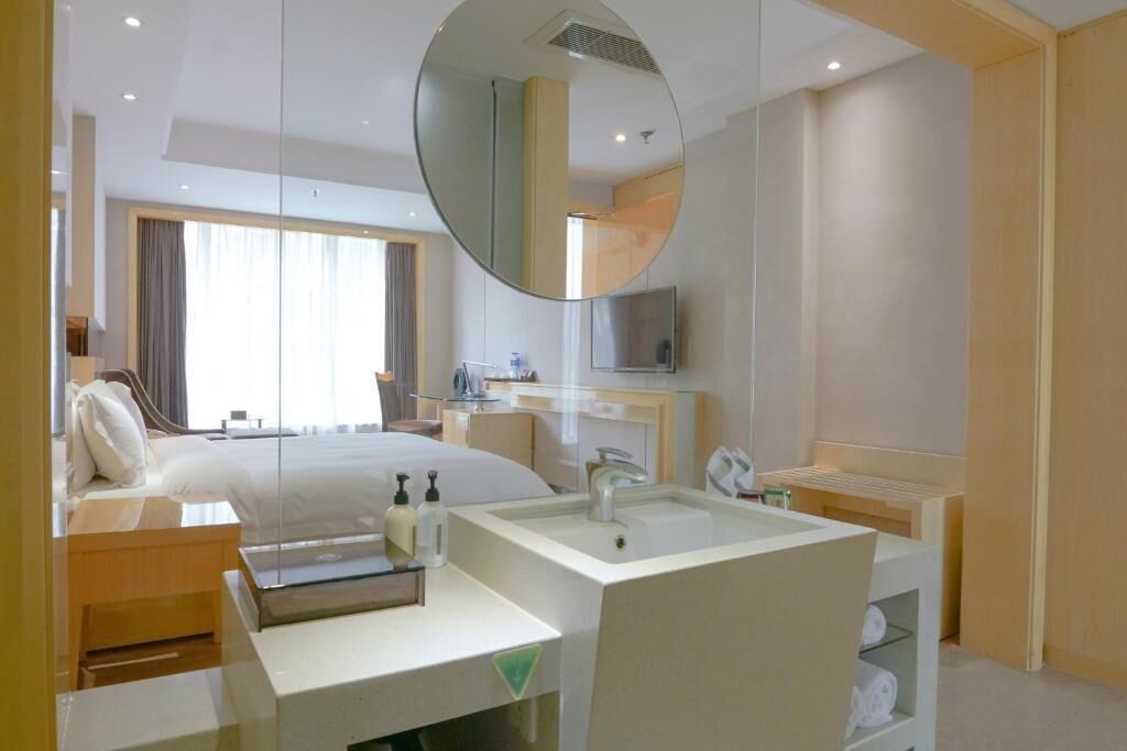 Quarto de hotel com cama, espelho grande, televisão, banheiro com pia e janela.