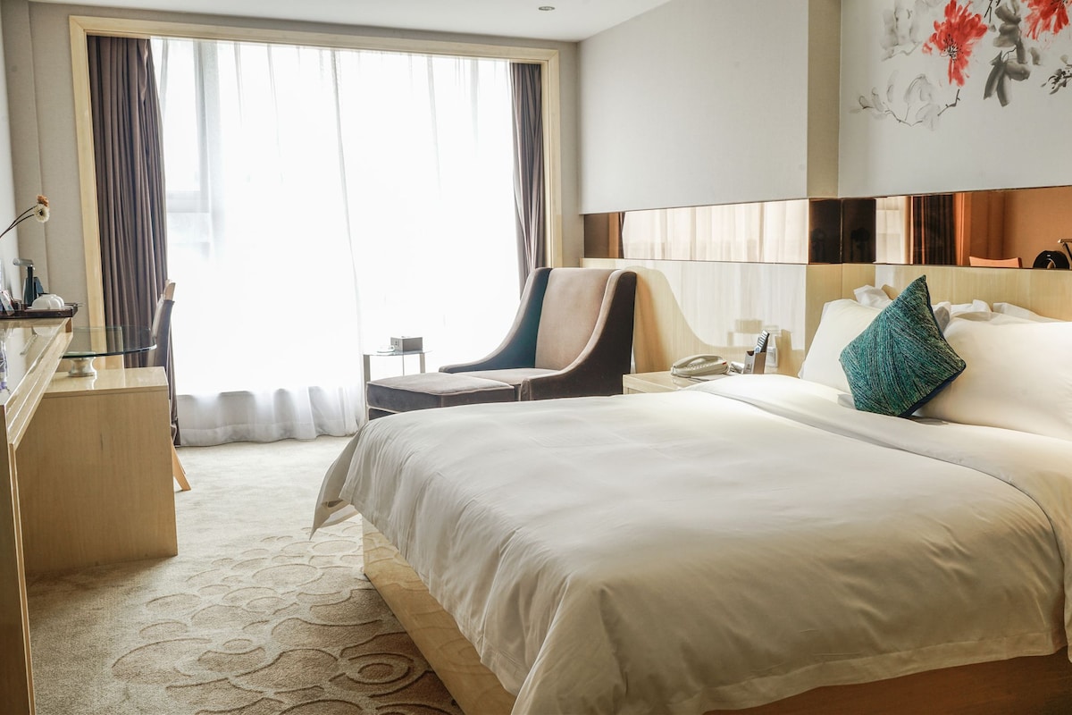 Quarto de hotel com uma cama grande, uma escrivaninha com cadeira, mesa de cabeceira e janela com cortinas.