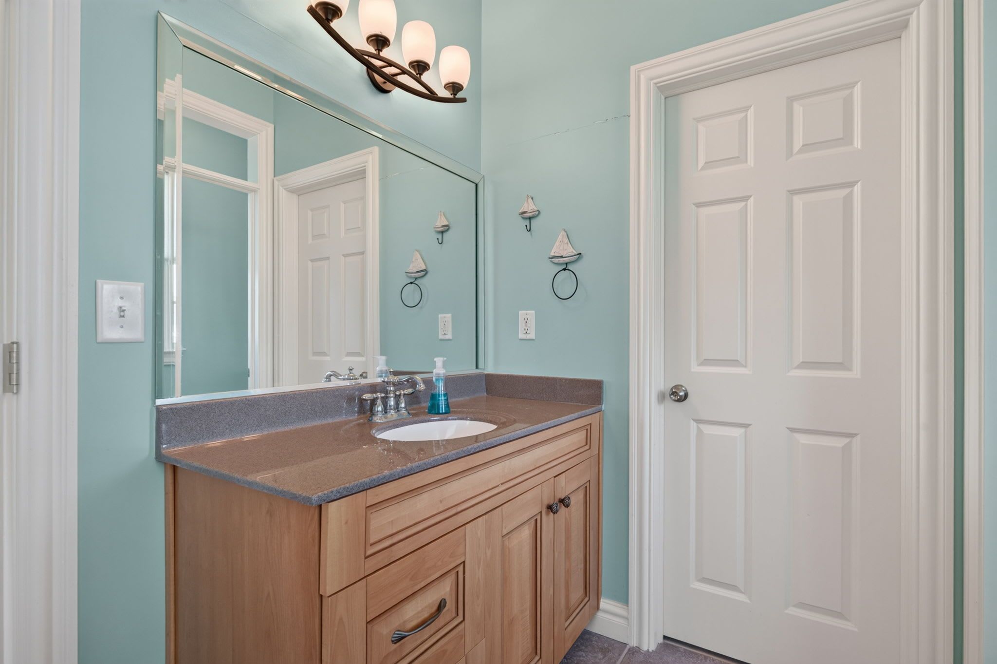 Une salle de bain avec un meuble-lavabo en bois, un évier et un miroir. Il y a une porte sur la droite et un luminaire au-dessus du miroir.