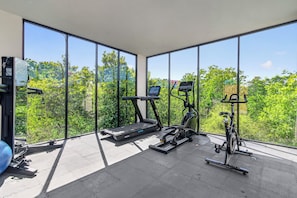 Academia com equipamentos de ginástica e amplas janelas com vista para uma paisagem verde.