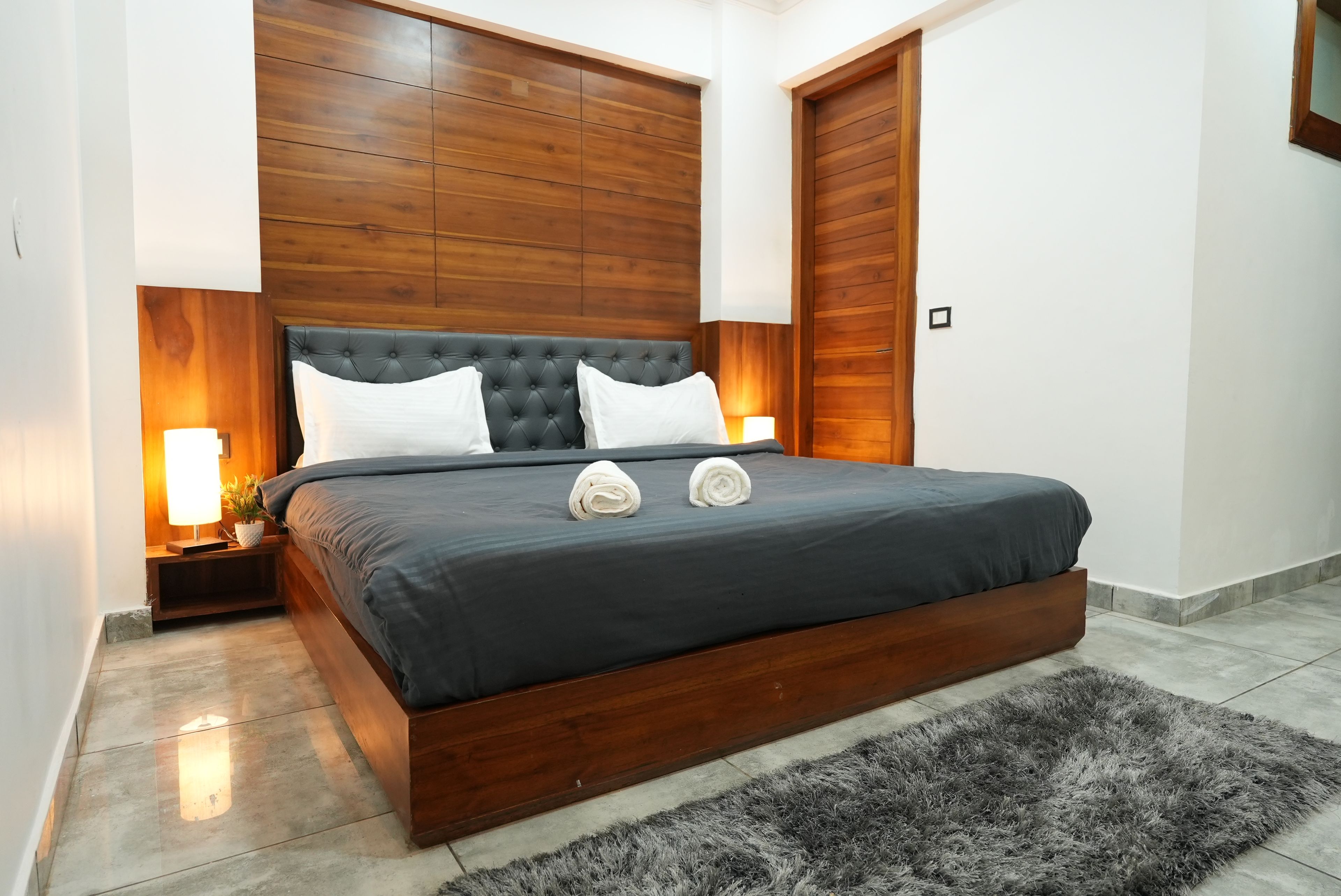 Un dormitorio moderno con cabecera de madera, dos lámparas de noche, una alfombra gris y una cama bien hecha con dos almohadas blancas.