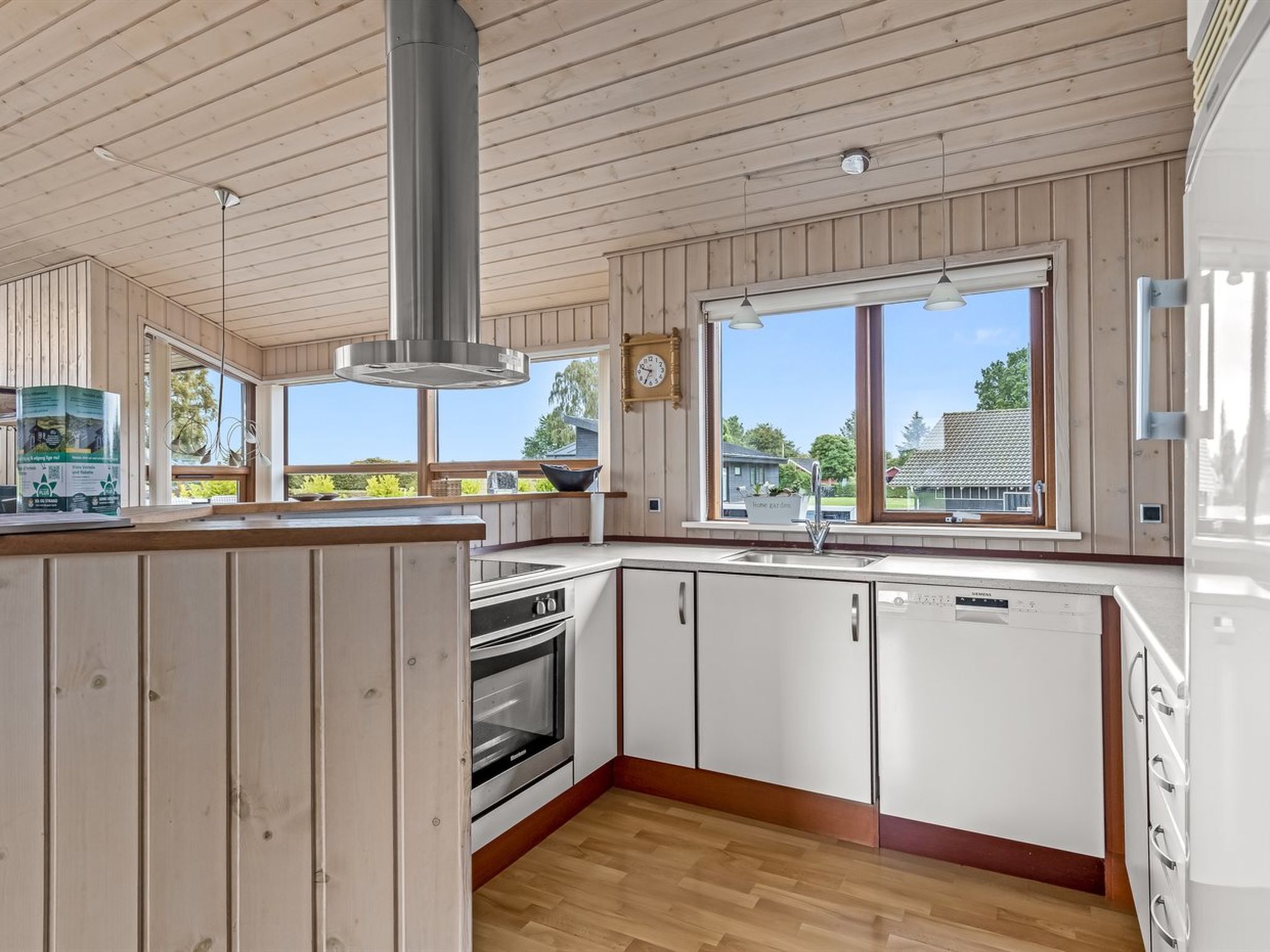 A modern kitchen with wooden paneling, a stainless steel range hood, a dishwasher, and a large window offering a view of the outdoors.