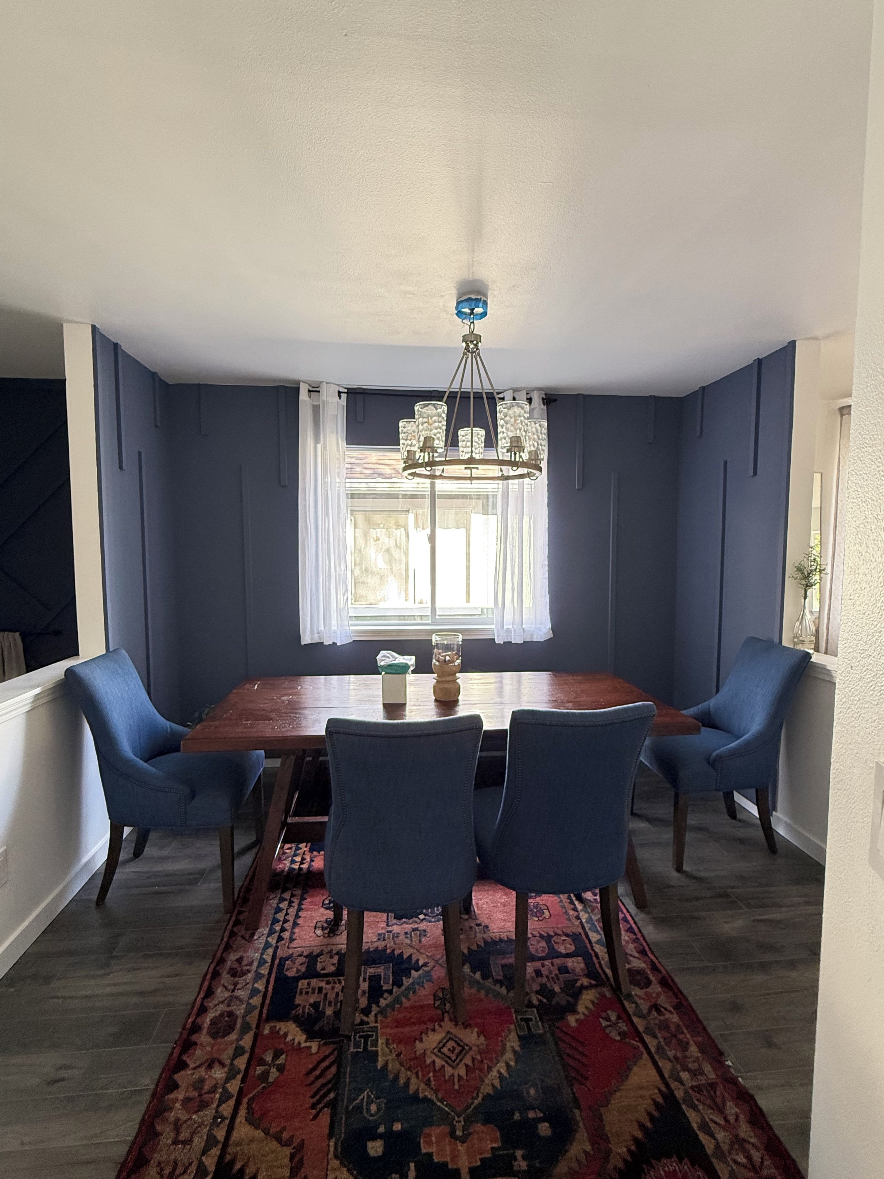 Une salle à manger avec une table en bois, des chaises bleues, un tapis à motifs et un lustre.
