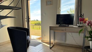 A modern living room with a TV, a desk, and a view of a green landscape through the window. - Charming Normandy Escape for Three (Courtils)