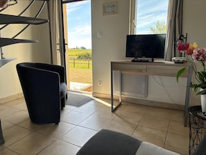 A modern living room with a TV, a desk, and a view of a green landscape through the window. - Charming Normandy Escape for Three (Courtils)