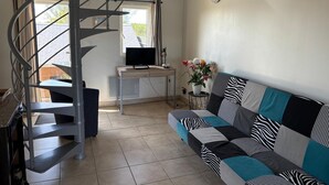 A modern living room with a spiral staircase, a flat-screen TV, a sofa with patterned cushions, and a small side table with a vase of flowers. - Charming Normandy Escape for Three (Courtils)